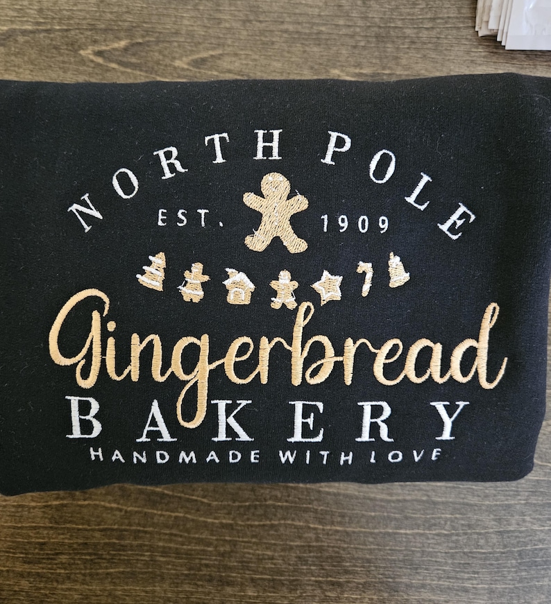May include: Black sweatshirt with white embroidered text that reads "North Pole Gingerbread Bakery Est. 1909 Handmade with Love". The text is surrounded by a gingerbread man and gingerbread cookies.
