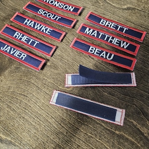 May include: A selection of navy blue and red name patches with white embroidered text. Names include BRONSON, SCOUT, HAWKE, RHETT, JAVIER, BRETT, MATTHEW, and BEAU. The patches have a hook and loop backing.
