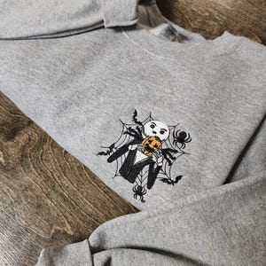 May include: A gray sweatshirt featuring an embroidered design of a skeleton character holding a pumpkin, surrounded by a spider web, spiders, and bats. The design is in black and orange.