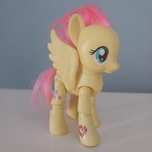 Op de afbeelding: Een gele speelgoedpony met roze haar en vleugels. De pony heeft blauwe ogen en roze markeringen op de benen. Het speelgoed staat op een wit oppervlak tegen een lichtblauwe achtergrond.