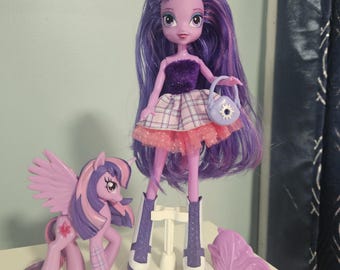 May include: A doll and pony set featuring a purple-haired doll with a sparkly top, plaid skirt, and a matching purse. A purple pony with wings and a tail is also included, along with a purple hair accessory. The set is on a white surface.