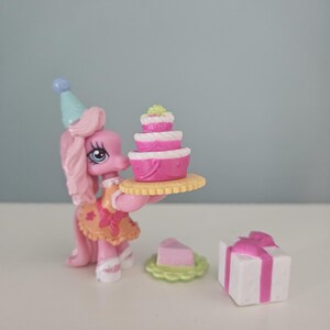 May include: A pink toy pony wearing a party hat and holding a tiered cake on a yellow plate. A slice of cake on a green plate and a white gift box with a pink ribbon are also present.