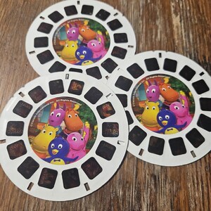 Puede incluir: Tres discos circulares blancos de View-Master con los personajes de The Backyardigans. Cada disco tiene una imagen central del grupo animado y varios fotogramas rectangulares alrededor del borde. El texto Viacom International Inc. All Rights Reserved está impreso en la imagen central.