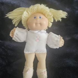 Könnte beinhalten: Vintage Cabbage Patch Kid Puppe mit hellgelber Garnfrisur. Die Puppe hat ein rundes, blasses Gesicht mit blauen Augen und einem kleinen, lächelnden Mund. Sie trägt ein weißes Kleid mit einer rosa Blume und beige Socken und Schuhen.