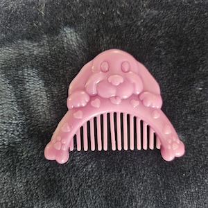 Puede incluir: Un peine de plástico rosa con una cabeza de perro moldeada en la parte superior. El mango curvo está decorado con pequeños corazones y huellas de patas en relieve, mientras que los dientes del peine se extienden desde la base del perro.