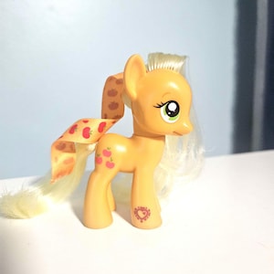 Può includere: Un pony giocattolo giallo con criniera e coda bionde e un nastro con disegni di mele rosse. Il pony ha un emblema a forma di cuore sulla zampa posteriore e occhi verdi. Il giocattolo è su una superficie bianca.