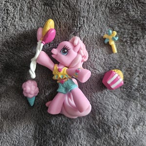 May include: A pink My Little Pony figurine with blue eyes, wearing a yellow top and blue shorts, holding a balloon-like object. Accessories include a cotton candy cone, a star-shaped wand, and a cake slice.