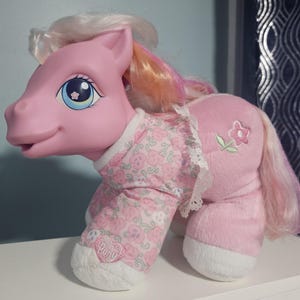 Könnte beinhalten: Ein rosa Plüschpferd mit weißer und rosa Mähne und Schweif. Das Pony trägt ein rosa Outfit mit Blumenmuster und weißer Spitzenborte. Auf der Seite ist eine Blume gestickt und auf dem Fuß ein herzförmiges Logo.