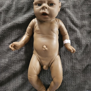 May include: A realistic, anatomically correct baby doll with brown skin tone. The doll is lying on a gray surface, with arms and legs extended. It has a detailed face with open eyes and a slight frown. A hospital bracelet is on its wrist.