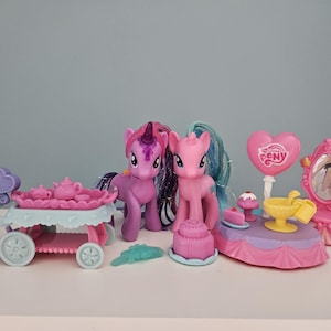 Op de afbeelding: Een verzameling My Little Pony-speelgoed, waaronder twee ponyfiguren, een theekar, een taart, een hartvormige ballon en een make-upspiegel. Het speelgoed is voornamelijk roze, paars en blauw.