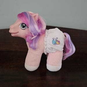 May include: Pink plush pony toy with purple and pink hair. The pony is wearing a white nappy with a colourful seahorse design.