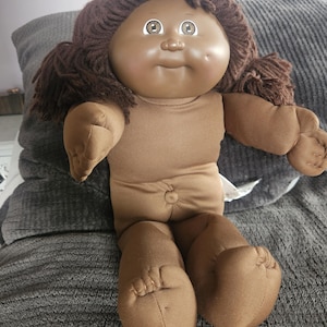 Peut inclure: Poupée vintage avec un teint brun, des cheveux en fil brun et de grands yeux expressifs. Le corps et les membres de la poupée sont en tissu beige. La poupée est assise sur une surface grise. Les traits de la poupée sont doux et arrondis.