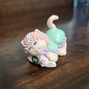 Può includere: Piccola statuina decorativa di un carlino in costume da gatto. Il carlino è rosa chiaro con un vestito verde e un colletto lavanda. La statuina ha occhi blu e un'etichetta con la scritta "Pug".