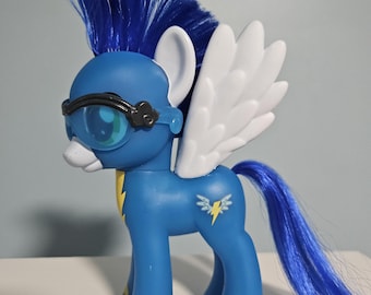 Op de afbeelding: Een blauw speelgoedpony met een blauwe hanenkam, witte vleugels en een lange blauwe staart. De pony heeft een bril en gele bliksemmarkeringen. Een wit bliksemontwerp staat op de flank van de pony.