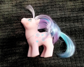 May include: A pink toy pony with a long, flowing mane and tail in shades of blue and purple. The pony has blue floral designs on its side and a white forelock. It is a vintage toy.