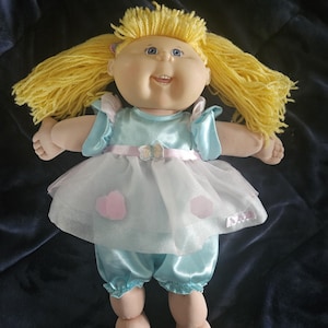 May include: A vintage Cabbage Patch Kid doll with yellow yarn hair and blue eyes. The doll is dressed in a light blue satin outfit with a white tulle skirt and pink heart accents. A pink bow is at the waist.