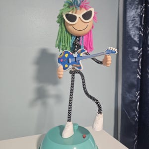 Peut inclure: Une figurine jouet fantaisiste avec un visage souriant, des lunettes de soleil et des cheveux en fil colorés. Elle tient une guitare bleue avec un motif arc-en-ciel. La figurine a des membres rayés noirs et blancs et des chaussures blanches, sur une base turquoise.
