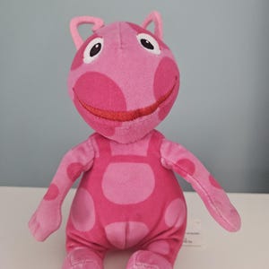 Puede incluir: Un peluche rosa de un personaje de dibujos animados con grandes lunares redondos. El personaje tiene una cara sonriente con labios rojos bordados y ojos grandes con bordes blancos. Tiene orejas pequeñas y curvas y está sentado erguido.