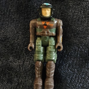 May include: A vintage action figure of a pilot in a brown and green uniform with a black helmet. The figure has a light skin tone and is standing upright. The figure's chest has a yellow and orange symbol.
