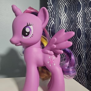 Puede incluir: Una figura de poni de plástico morado con cuerno y alas, que luce una larga melena multicolor en tonos morado, rosa y amarillo. La figura tiene grandes ojos morados y una marca en forma de estrella en el costado con detalles en blanco.
