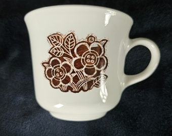 Op de afbeelding: Witte keramische koffiemok met een bruin bloemenontwerp. Het ontwerp bevat gestileerde bloemen en bladeren.
