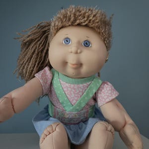 Puede incluir: Una muñeca vintage con cuerpo de tela suave y cabeza de vinilo. La muñeca tiene ojos azules, expresión sonriente y cabello castaño claro en cola de caballo. Viste un top estampado rosa y blanco, un peto verde y una falda azul.