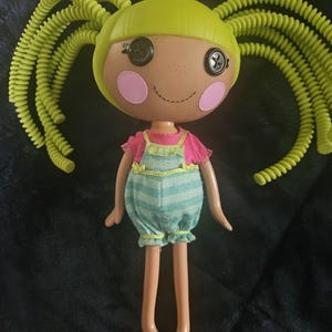 May include: A doll with button eyes, pink cheeks, and a stitched smile. The doll has lime green hair in pigtails, a pink top, and blue striped dungarees with yellow trim. The doll's legs are long and slender.
