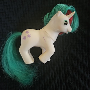 Puede incluir: Un juguete de pony My Little Pony unicornio blanco con una crin y cola de color verde azulado y rojo. El pony tiene marcas de hojas moradas en su flanco.