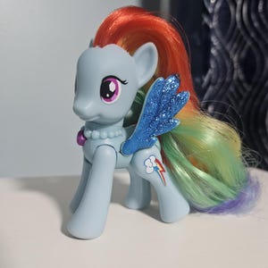 May include: A light blue pony figurine with a rainbow-colored mane and tail. The figure features glittery blue wings, a purple gem accent on the chest, and a cloud with a rainbow lightning bolt symbol printed on the rear flank. The eyes are large and purple.