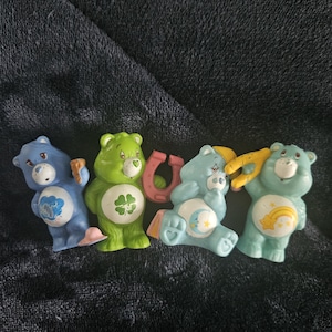 May include: Four Care Bear figurines in various colors. A blue bear holds a cloud, a green bear has a four-leaf clover, a light blue bear has a moon, and a light blue bear has a rainbow. A pink horseshoe is also present.