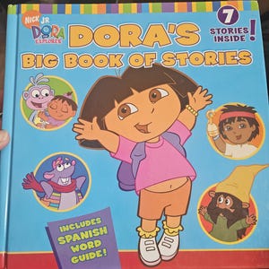 Op de afbeelding: Een kinderboek getiteld "Dora's Big Book of Stories" met Dora the Explorer op de omslag. Het boek is blauw met kleurrijke illustraties en tekst, waaronder "Includes Spanish Word Guide!" en "7 Stories Inside!"