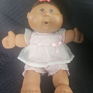 May include: A vintage doll with brown skin, brown hair in a bun with a pink bow, and large brown eyes. The doll is dressed in a pink floral print dress with a white overlay and matching shorts. The doll's arms are outstretched.