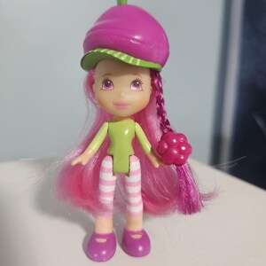 Peut inclure: Une petite poupée avec des cheveux roses et un chapeau en forme de baie. La poupée a un haut vert, des jambes rayées blanches et roses et des chaussures violettes. Elle tient une framboise rose. Le chapeau est magenta avec un bord vert.