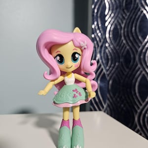 May include: A small doll with yellow skin, large blue eyes, and long wavy pink hair featuring a small white butterfly accessory. The doll wears a white sleeveless top, a mint green skirt with pink butterfly patterns, and tall mint green boots.