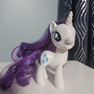 May include: A white plastic unicorn figurine with a spiraled horn, large blue eyes, and long, flowing purple and dark blue synthetic hair. The flank features a cutie mark consisting of three light blue, four-pointed star-like symbols.