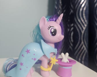 May include: A light purple unicorn figurine with a blue and purple mane, wearing a light blue cape with pink star patterns. The unicorn holds a yellow star-tipped wand and stands next to a purple top hat containing a small white rabbit figure.