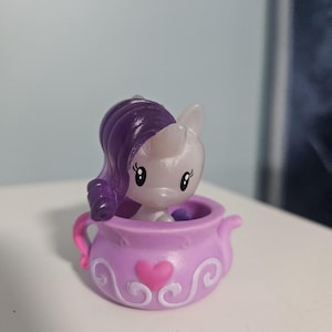 Puede incluir: Pequeña figura morada de un pony con cuerpo blanco y pelo morado, sentado dentro de un recipiente con forma de tetera lavanda. El recipiente tiene un corazón rosa y espirales blancas. El pony tiene ojos grandes y un cuerno.