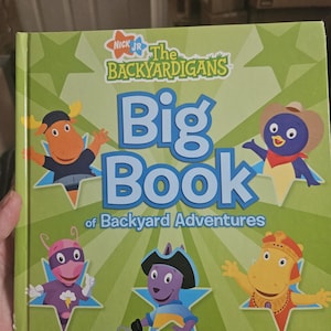 Puede incluir: Un libro infantil verde titulado "Big Book of Backyard Adventures" con personajes del programa Nick Jr., The Backyardigans. La portada tiene acentos de estrellas e ilustraciones de personajes.