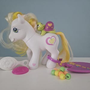 May include: A white toy pony with yellow and blonde hair, a green turtle figurine, and accessories. The pony has a heart-shaped design on its side. Additional items include a brush, a pink disc, a purple surfboard, and colourful beads.