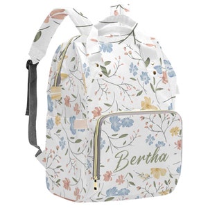 May include: A white diaper bag with a floral pattern in shades of blue, pink, and yellow. The bag has a zippered pocket on the front with the name "Bertha" written in green cursive.