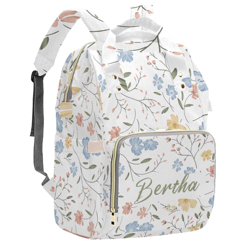 May include: A white diaper bag with a floral pattern in shades of blue, pink, and yellow. The bag has a zippered pocket on the front with the name "Bertha" written in green cursive.