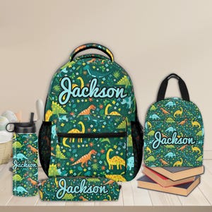 May include: A green backpack, lunch bag, pencil case, and water bottle with a dinosaur pattern and the name "Jackson" on each.