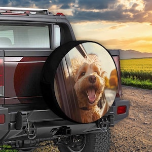 May include: A black spare tire cover with a photo of a light brown dog looking out of a car window. The dog is smiling and has its tongue sticking out. The background is a blurry image of a road and a field.