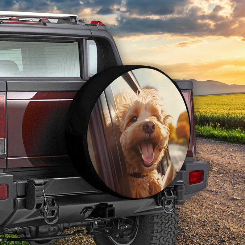 May include: A black spare tire cover with a photo of a light brown dog looking out of a car window. The dog is smiling and has its tongue sticking out. The background is a blurry image of a road and a field.