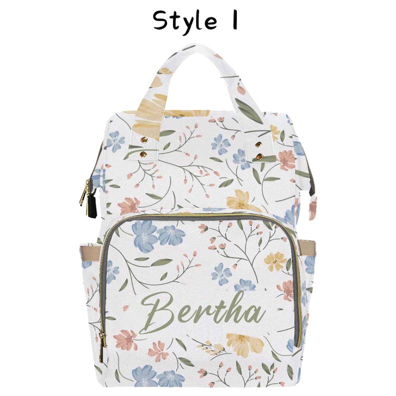 May include: White diaper bag with a floral pattern and the name Bertha printed in green on the front. The bag has a top handle and adjustable straps.