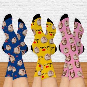 May include: Three pairs of colorful socks with a repeated pattern of a child's face and the text "I love mama" in black letters with a red heart. The socks are blue, yellow, and pink.