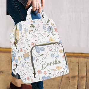 May include: A white floral print diaper bag with a zippered pocket and the name "Bertha" written in cursive on the front.