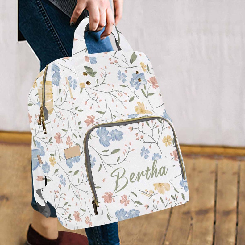 May include: A white floral print diaper bag with a zippered pocket and the name "Bertha" written in cursive on the front.