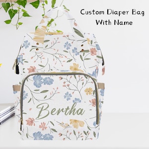 May include: White diaper bag with a floral pattern in shades of blue, yellow, and pink. The bag has a zippered pocket on the front and the name "Bertha" is embroidered in green.