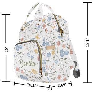May include: A white diaper bag with a floral pattern in shades of blue, pink, and yellow. The bag has a large main compartment, a front pocket with the name "Bertha" embroidered on it, and adjustable straps. The bag measures 18.1 inches tall, 10.83 inches wide, and 6.69 inches deep.